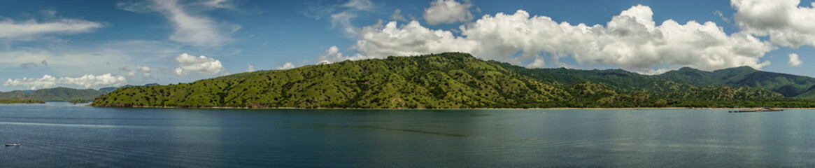 Fototapeta premium Komodo Island, Indonesia - February 24, 2019: Panorama shot of Komodo National Park in blue sea. Green forested hills in back under blue sky with white clouds. Strip of sandy beach with pier.