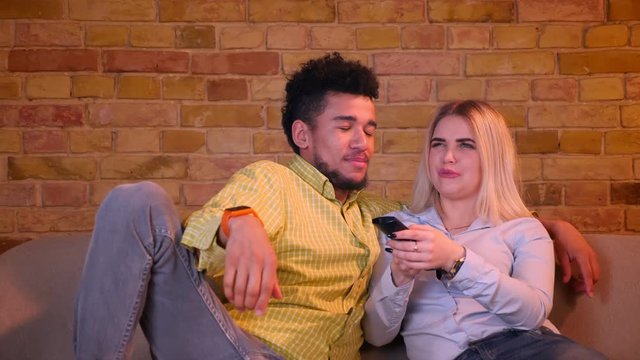 Multinational couple choosing programes with remote controller watching TV in cozy home atmosphere.