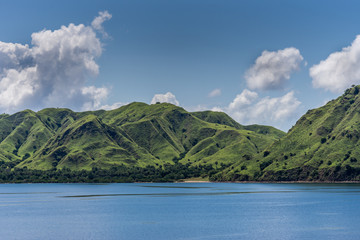 Komodo Island, Indonesia - February 24, 2019: Green mountain range descending on sand beach under blue sky with cloudscape, part of Komodo National Park.