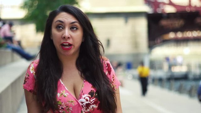  Young Latina woman  talking head is at Chicago River Walk  having an exciting conversation and happy showing many facial expression while moving hair.  wearing pink blouse