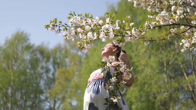 Young traveler pregnant woman enjoys her leisure free time in a park with blossoming sakura cherry trees wearing a summer light long dress with flower pattern - European baltic city Riga, Latvia
