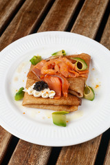 Russian crepes with smoked salmon, black caviar, avocado and cream cheese for shrovetide on white dish and wooden table
