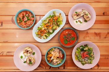 Top view of various vegetarian meals, served on a light wooden table: poke bowl, gazpacho, vegetable salads, BBQ fruits and vegan noodle soup