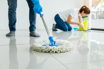 Young housekeeper cleaning floor mobbing holding mop and plastic bucket with brushes, gloves and detergents in the leaving room house floor helping his wife