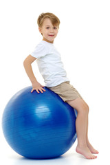 The little boy on the big ball is engaged in fitness. White T-sh