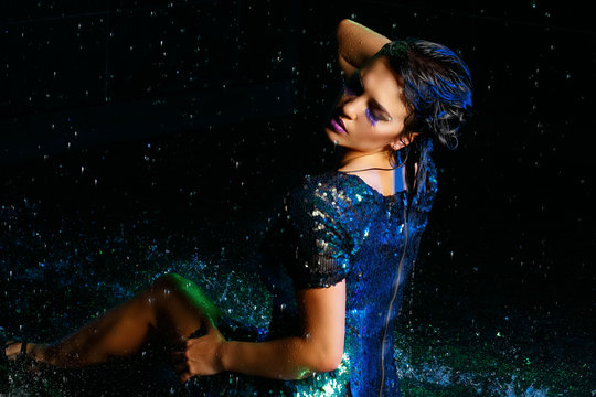 Young sexy woman sitting on wet floor with water drops. Gren light effect. Seductive girl dressed in a sparkling dress in sequins resembling fish scales in a dark room. Water studio, spray, splashes.