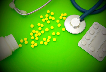 Stethoscope, blister with white pills, inverted jar of vitamins With yellow, scattered on a green...