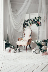 vintage armchair decorated with flowers and greens, stands in a classic room on a white wooden floor surrounded by lighted candles near  large window and curtains