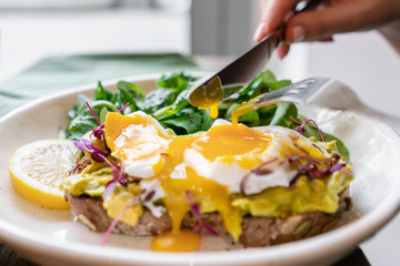 Young woman eat breakfast with knife and fork, Morning in cafe. Healthy breakfast with wholemeal bread toast with avocado, poached egg with green salad