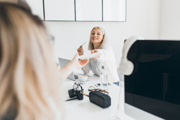 Naklejka premium Smiling blonde secretary handed documents to colleague and drinking coffee. Fair-haired female programmer posing near computer with black screen while her friend enjoying latte.