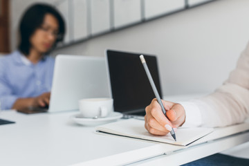 Photo of female hand with white manicure holding pencil with asian office worker on background. Students writing reports before university conference.