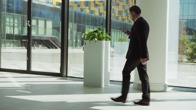 Side view tracking left shot of middle aged manager in black suit text messaging on smartphone while walking along entrance hall in modern business center and finally entering door