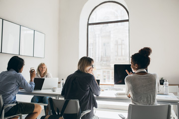 Indoor portrait from back of office worker sitting at the table and waiting chief before conference. Asian young man in blue shirt talking with blonde girl while using laptop in room with big windows.