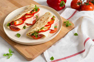 Italian piadina romagnola flatbread with red pepper, tomatoes, prosciutto ham, cheese and basil on the plate on white wooden background. Copy space