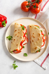 freshly baked italian piadina with cheese, tomato, ham, red pepper and basilio on a white plate on a white table. Top view, flat lay.