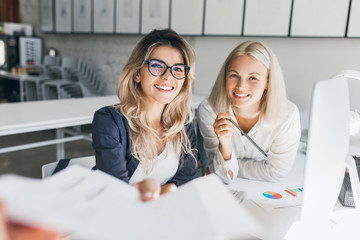 Indoor portrait of smiling blonde girls working together in office. Pretty woman in glasses spending time with friend during break and posing for photo in library. - obrazy, fototapety, plakaty