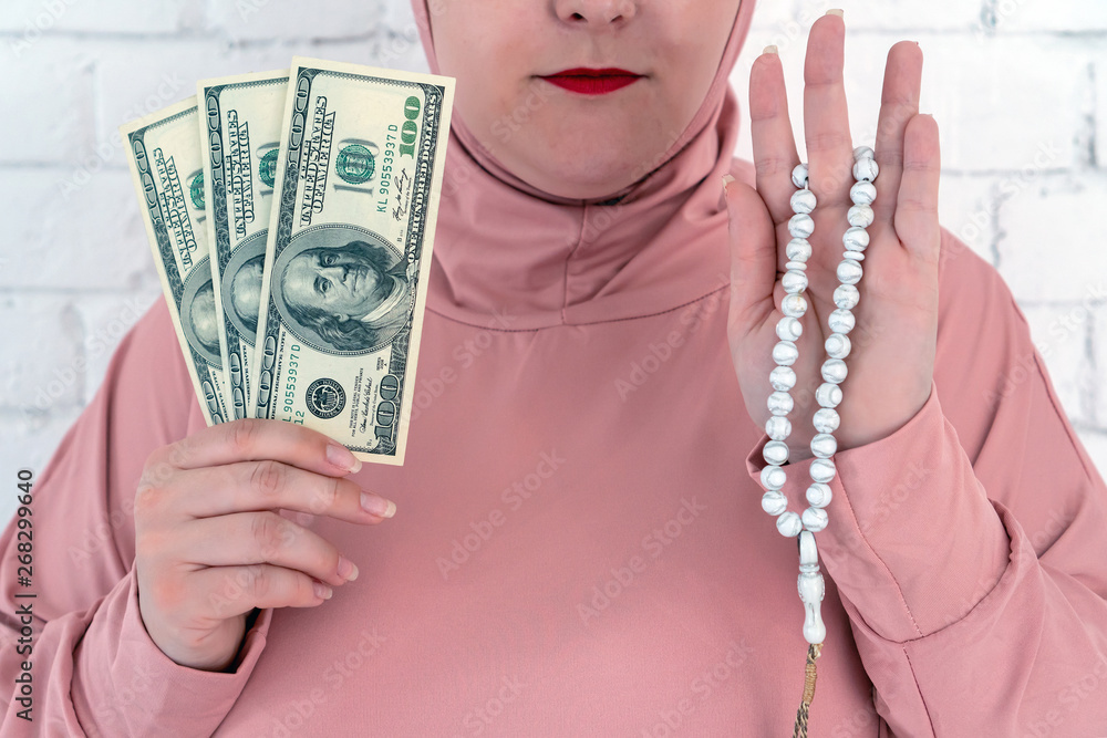 Wall mural White woman with blue eyes in a pink hijab holding a rosary and dollars on a white background.