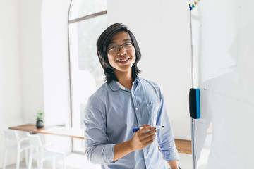 Confident japanese student in trendy glasses holding marker, standing near white board. Asian...
