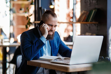 Serious bearded businessman conducts telephone conversations on a mobile phone at the computer. Unhappy man scolds on the phone. Emitations of rage in the office at the manager