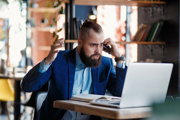 Obraz premium Serious bearded businessman conducts telephone conversations on a mobile phone at the computer. Unhappy man scolds on the phone. Emitations of rage in the office at the manager
