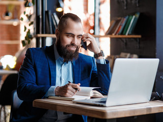 Obraz premium young bearded businessman at work in the office. A man sits at a laptop talking through a mobile phone. telephone conversations in coworking