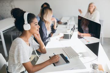 Charming black web-developer listening music in big headphones and using tablet. Indoor portrait of...