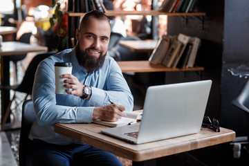 Fototapeta na wymiar Young serious bearded businessman working on computer at table,drinking coffee.Man analyzes information, data, develops business plan. Freelancer, entrepreneur.Online marketing, education, e-learning