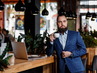 Young serious bearded businessman working on computer at table,drinking coffee.Man analyzes information, data, develops business plan. Freelancer, entrepreneur.Online marketing, education, e-learning