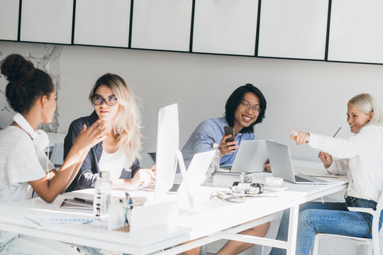 Asian laughing man making selfie with blonde girl in office while charming ladies beside talking with each other. Indoor portrait of young colleagues having fun together during workday.