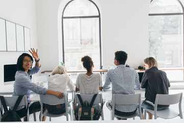 Portrait from back of african students having lunch between lectures. Smiling asian freelance worker looking over shoulder and waving hand while his friends talking.
