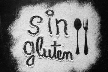 Gluten free flour with text gluten free in Spanish language with spoon and fork silhouette made with flour on dark texture background