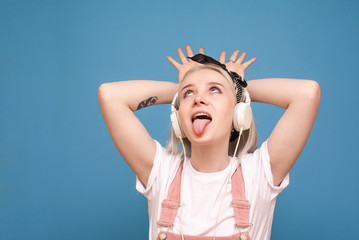 Cheerful girl in light clothing and headphones makes a funny face on a blue background. An emotional teen girl listening to music on a blue background.