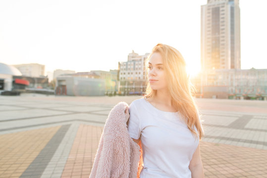 Street portrait of a girl who poses on a camera with a pink coat in her hand and looks sideways on the background of a beautiful sunset.Attractive girl at sunset