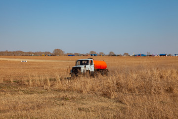 Naklejka premium A truck for the transportation of gasoline and fuel with an orange tank rides in a yellow field on the road during the delivery of fuel and lubricants to hard-to-reach regions and villages.