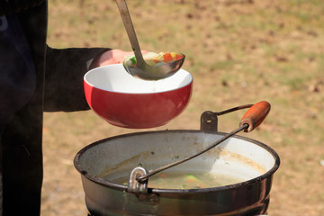 A person puts a fish soup cooked on a campfire from a bucket into a plate