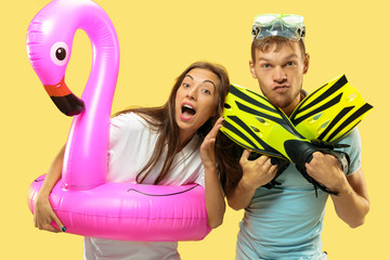 Beautiful young couple's half-length portrait on yellow studio background. Woman and man in caps and sunglasses standing with swimming rings and flippers. Facial expression, summer, weekend concept.
