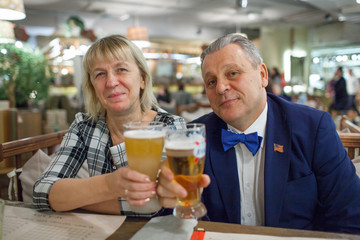 A portrait of a middle aged couple sitting at the table close to each other. Man is wearing a blue jacket and a bright blue bow tie. A woman is in a checkered shirt. Both of them are raising glasses