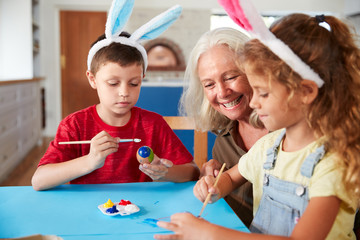 Grandmother With Grandchildren Wearing Rabbit Ears Decorating Easter Eggs At Home Together