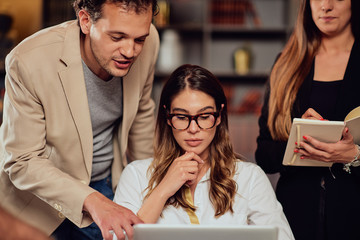 Fototapeta premium Charming buisnesswoman with brown hair and dressed elegant sitting at restaurant and looking at laptop. Next to her standing two colleagues and looking at laptop, too.