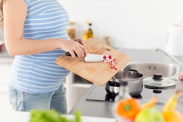 Obraz na płótnie Canvas pregnancy, cooking food and healthy eating concept - close up of pregnant woman with kitchen knife adding chopped raddish ftom wooden cutting board to pot on stove at home