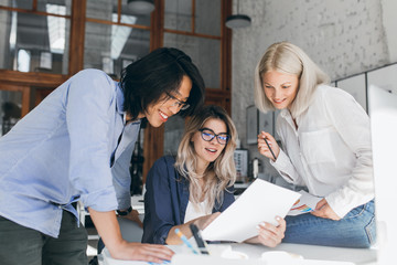 Fototapeta premium Beautiful blonde girl in glasses showing report to asian colleague which standing beside her table. Indoor portrait of cheerful international students analysing exam tasks.