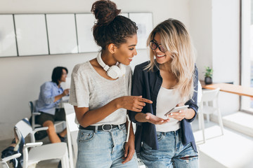 Blonde girl in jeans and glasses holding smartphone while talking with african friend in office....
