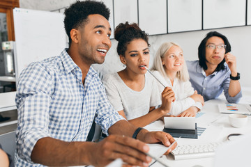 Pretty african female student biting pencil while thinking about something. Indoor portrait of pleased black office worker in checkered blue shirt sitting at the table with colleagues.