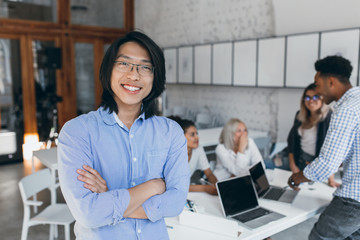 Excited asian young man in stylish glasses standing with arms crossed in library while his friends talking on background. Pleased foreign student passed all exams and happy for it.
