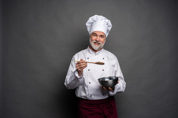 Smiling mature male chef with bowl and cooking vane in hands on black background.