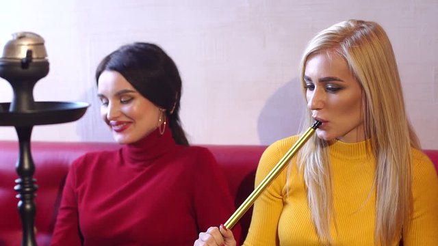 Two sexy girls smoke hookah in a cafe, they sit on a big red sofa and let out a lot of smoke from the mouth. A group of friends relax in a cafe, they talk, laugh and smoke a hookah. Slow motion.