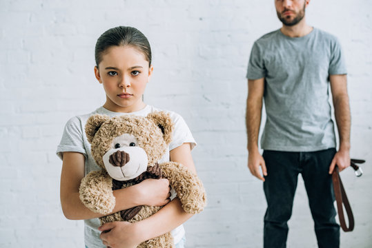 partial view of abusive father with belt and sad daughter holding teddy bear