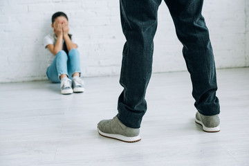 cropped view of father holding belt and scared daughter sitting on floor