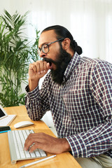 Fototapeta premium Bearded young manager yawning while typing on computer keyboard at office desk