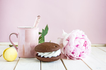 A pink tea mug on a saucer next to a brown cake with a white layer and a yellow macarons against the background of fresh yellow wildflowers and pink peony. Beautiful breakfast, Copy space.